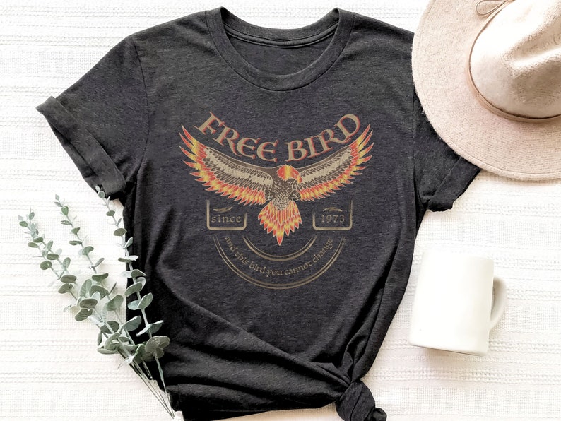 May include: A dark gray t-shirt with a vintage-style graphic of a bird with orange and yellow flames. The graphic includes the text 'Free Bird' and 'since 1973 and this bird you cannot change.'