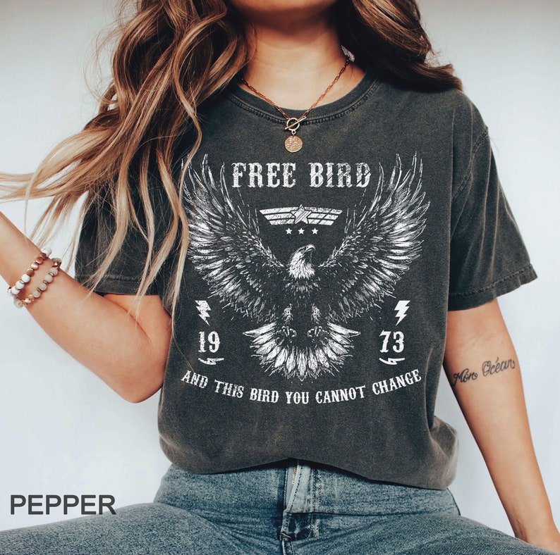 May include: A gray t-shirt with a white eagle graphic and the text 'FREE BIRD' above the eagle. The eagle has its wings spread and is flying. Below the eagle is the text 'AND THIS BIRD YOU CANNOT CHANGE' and the numbers '19' and '73' on either side of the eagle. The t-shirt is worn by a person with long brown hair.