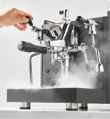 Hand adjusting a shiny espresso machine, releasing steam.